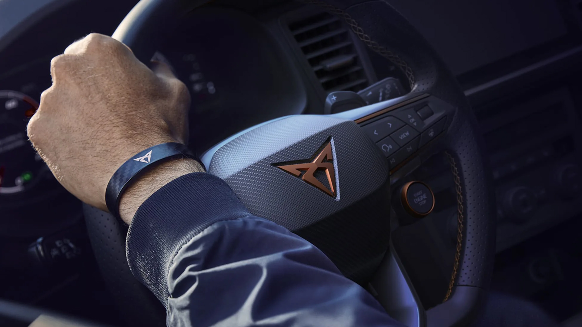 Detailaufnahme eines CUPRA-Lenkrads mit kupferfarbenem Logo, Hand am Steuer mit CUPRA-Armband